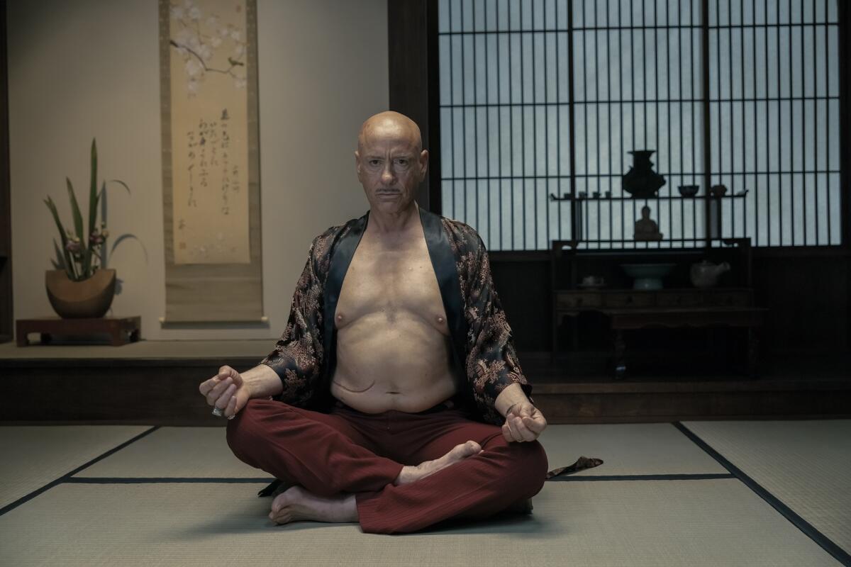 A man in a robe and pants sits with his legs crossed on the floor.