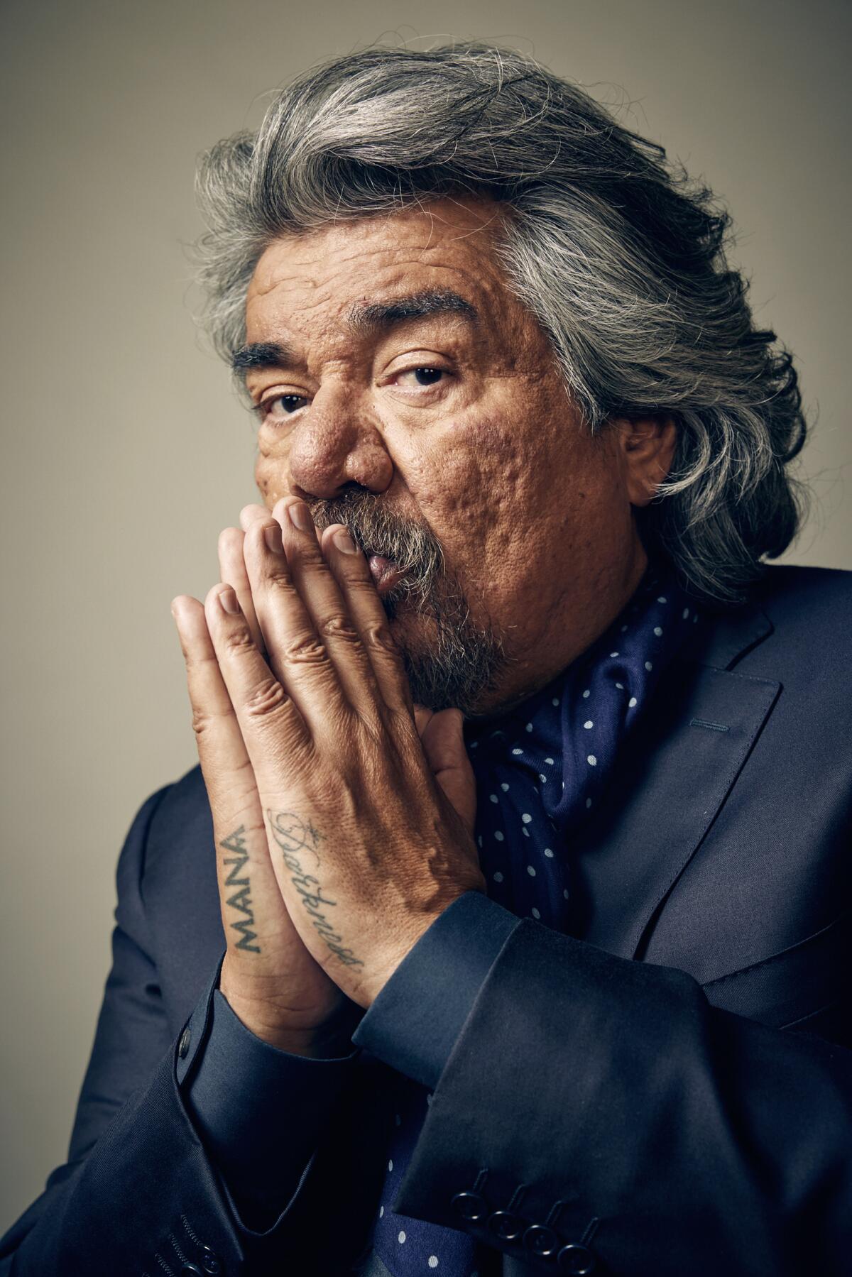 A vertical portrait of George Lopez holding his hands with the palms together in front of his mouth