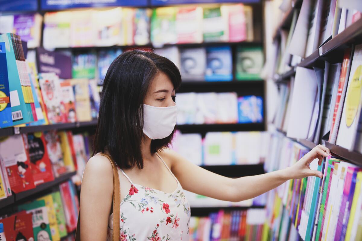 Asian woman wearing face mask choosing book magazine in book store. 