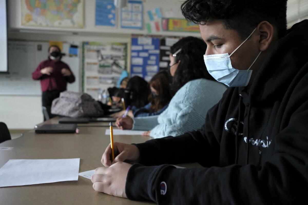 Students sit at desks and write with pencils. In the background is a teacher and a wall covered with notes and a map.