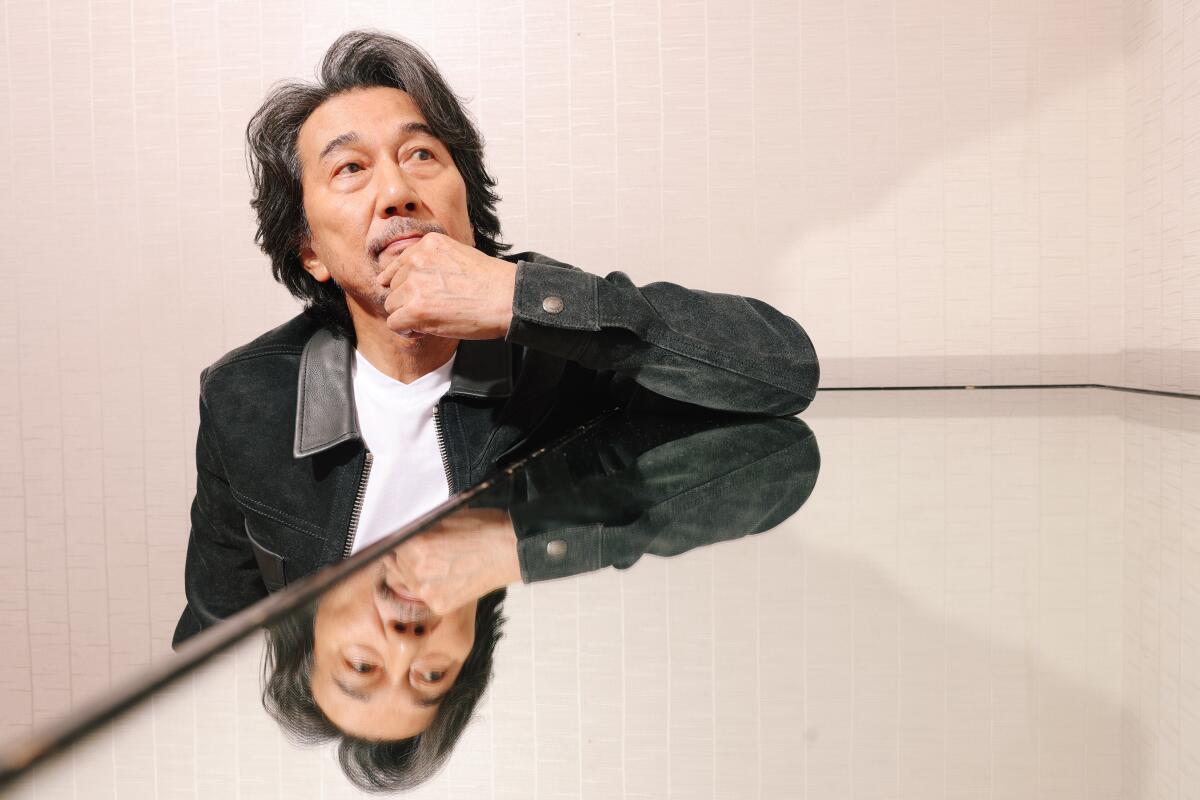 Koji Yakusho leans his elbow on a glass table, reflecting his face back to him, for a portrait. 
