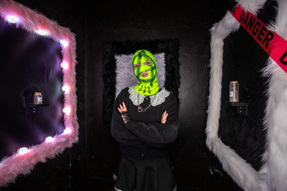 A woman wearing a neon green mask stands amid framed artworks.