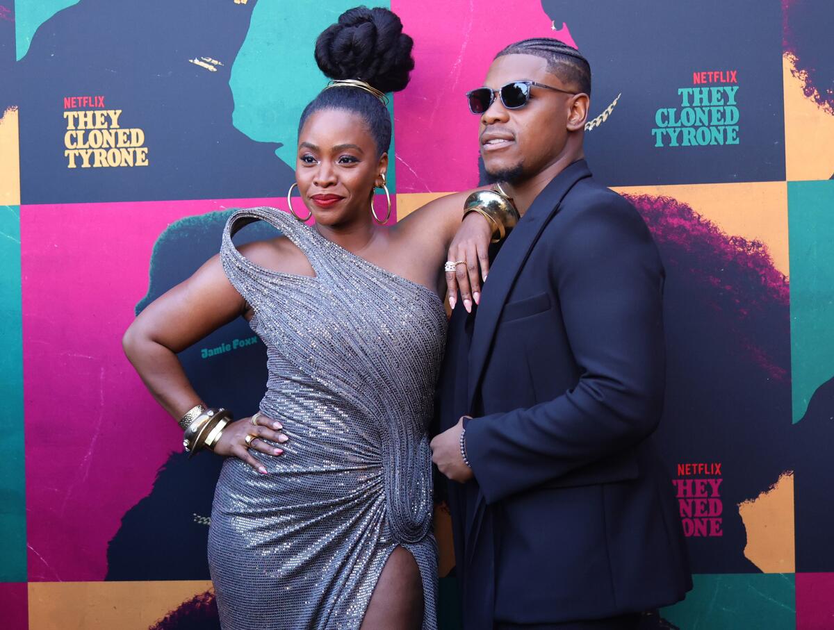 Teyonah Parris in a sparkly gown and John Boyega in a dark suit and wearing sunglasses stand in front of a backdrop