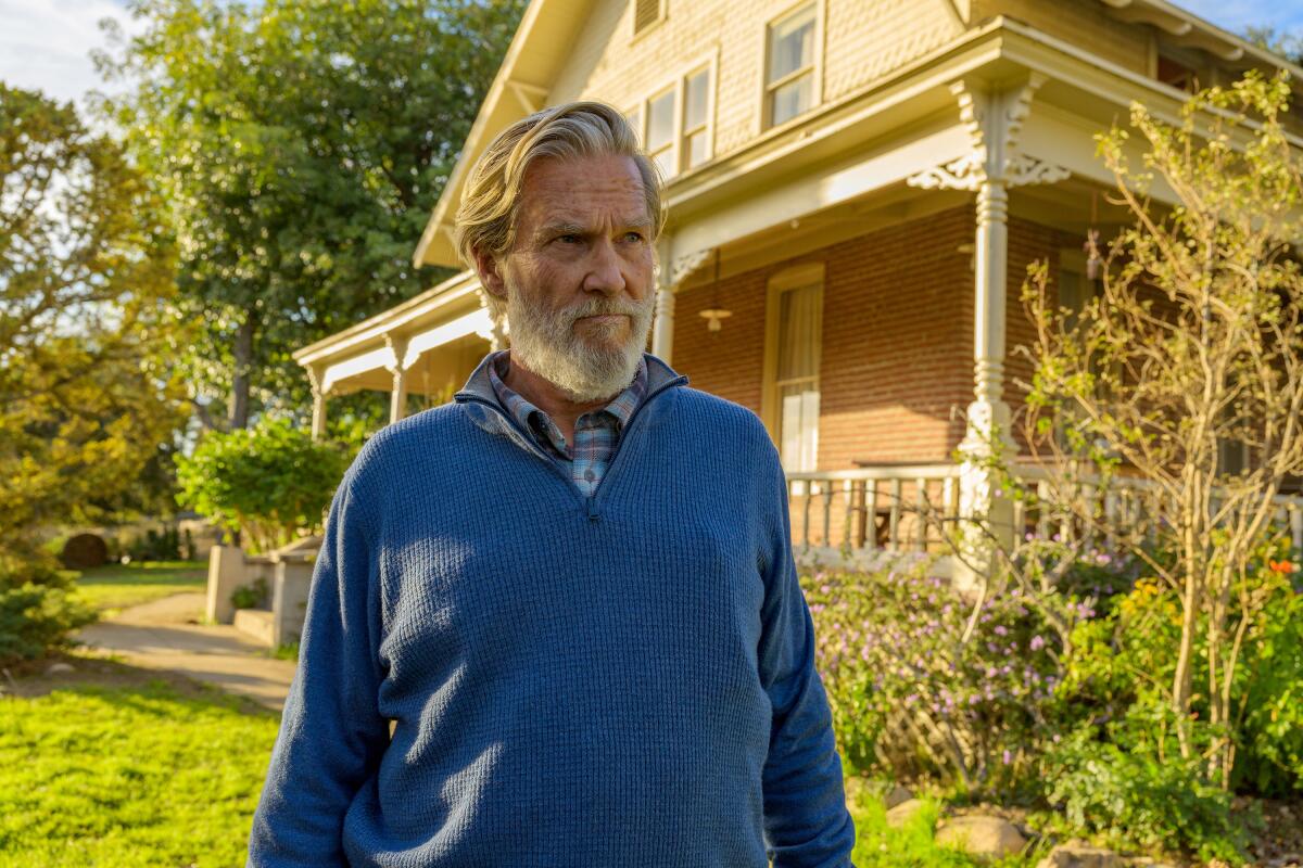 A bearded old man standing outside his bucolic Vermont home.
