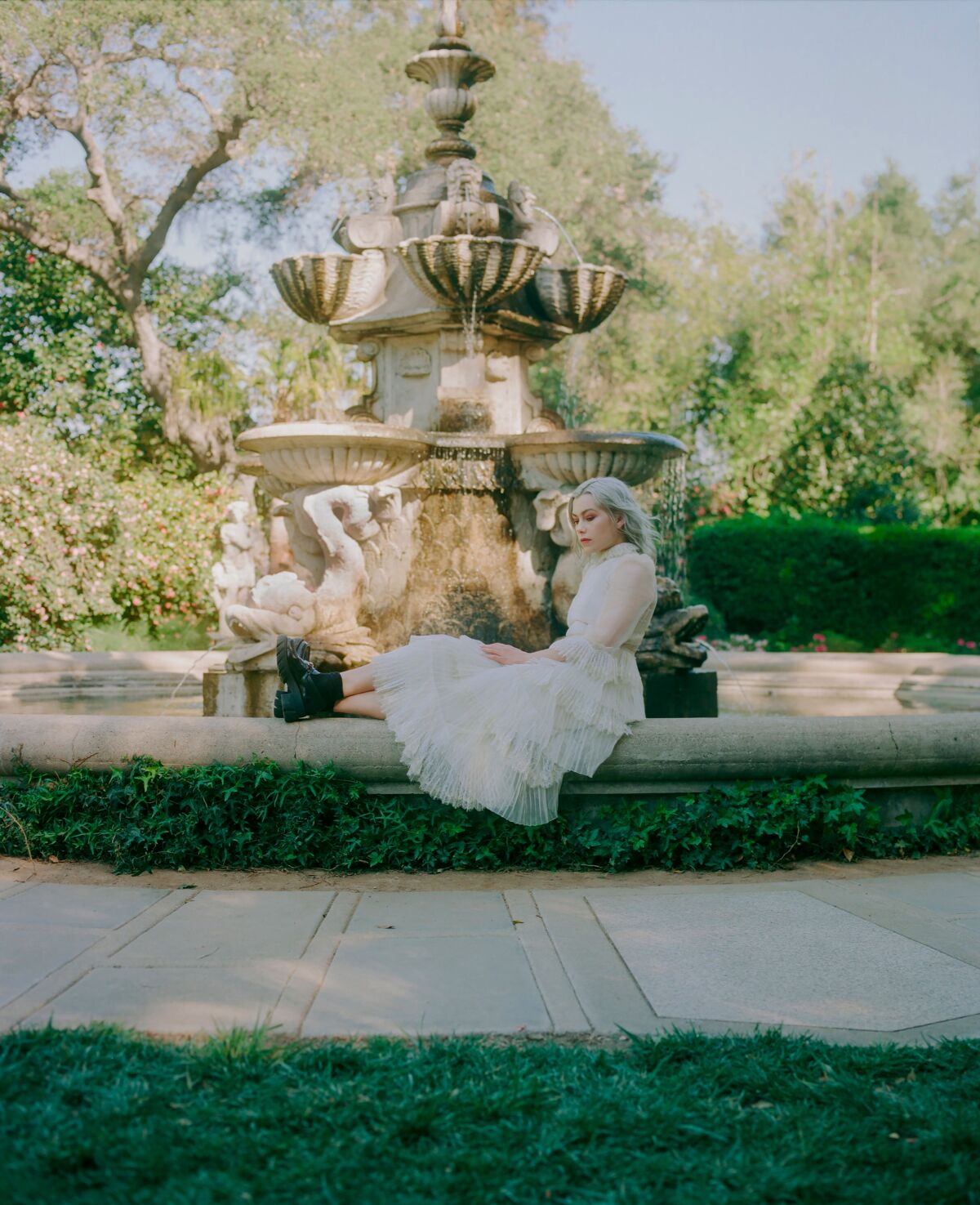 A woman in a white dress sits on the edge of a fountain in a garden.