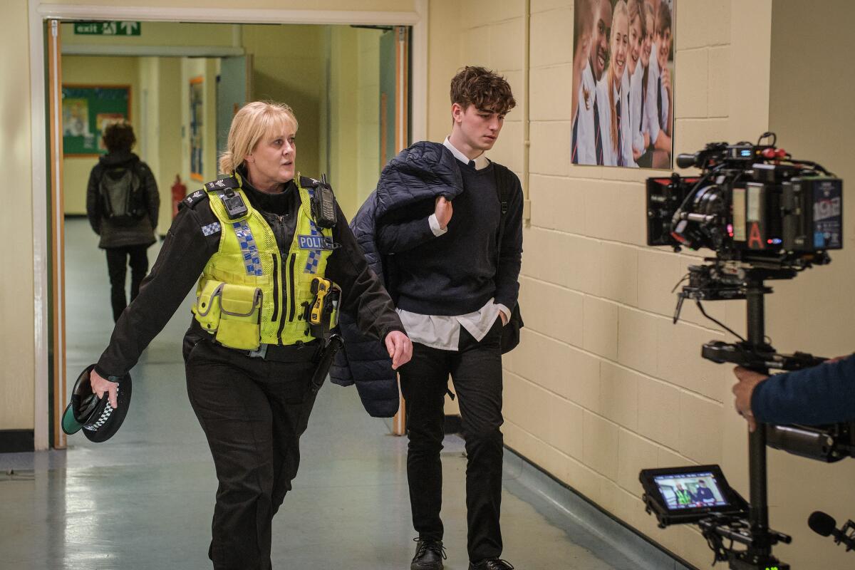 A woman dressed in a police vest and a teenage boy walk down a hallway, filmed by a camera.
