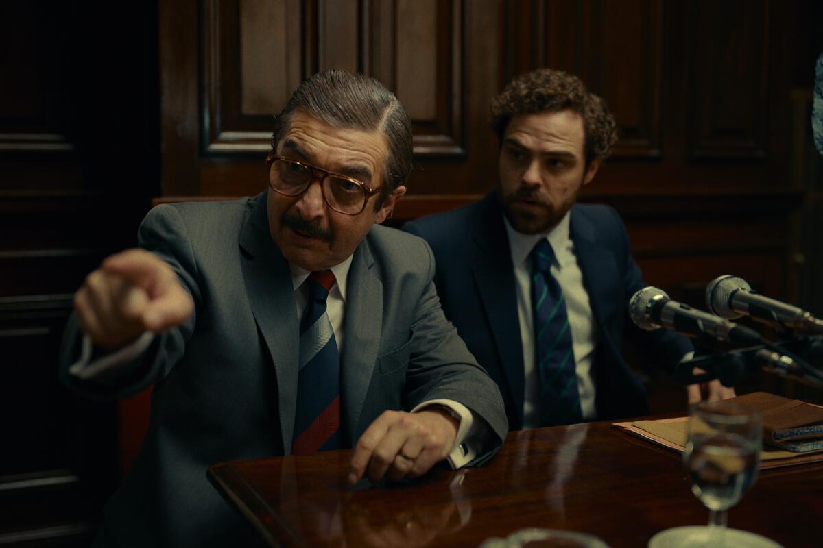 A man seated beside another man in a courtroom points off-camera in the movie "Argentina, 1985."