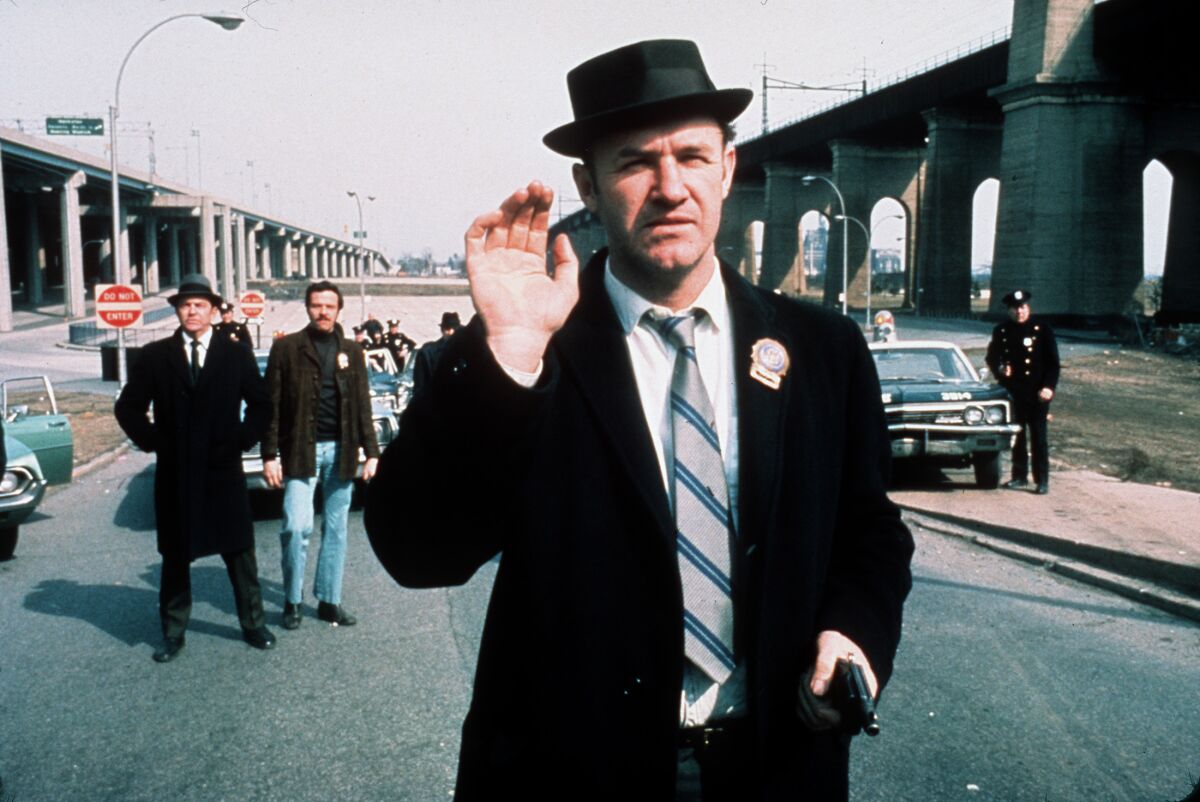 A detective wearing a porkpie hat holds one hand up and has a gun in the other as other officers stand by behind him. 