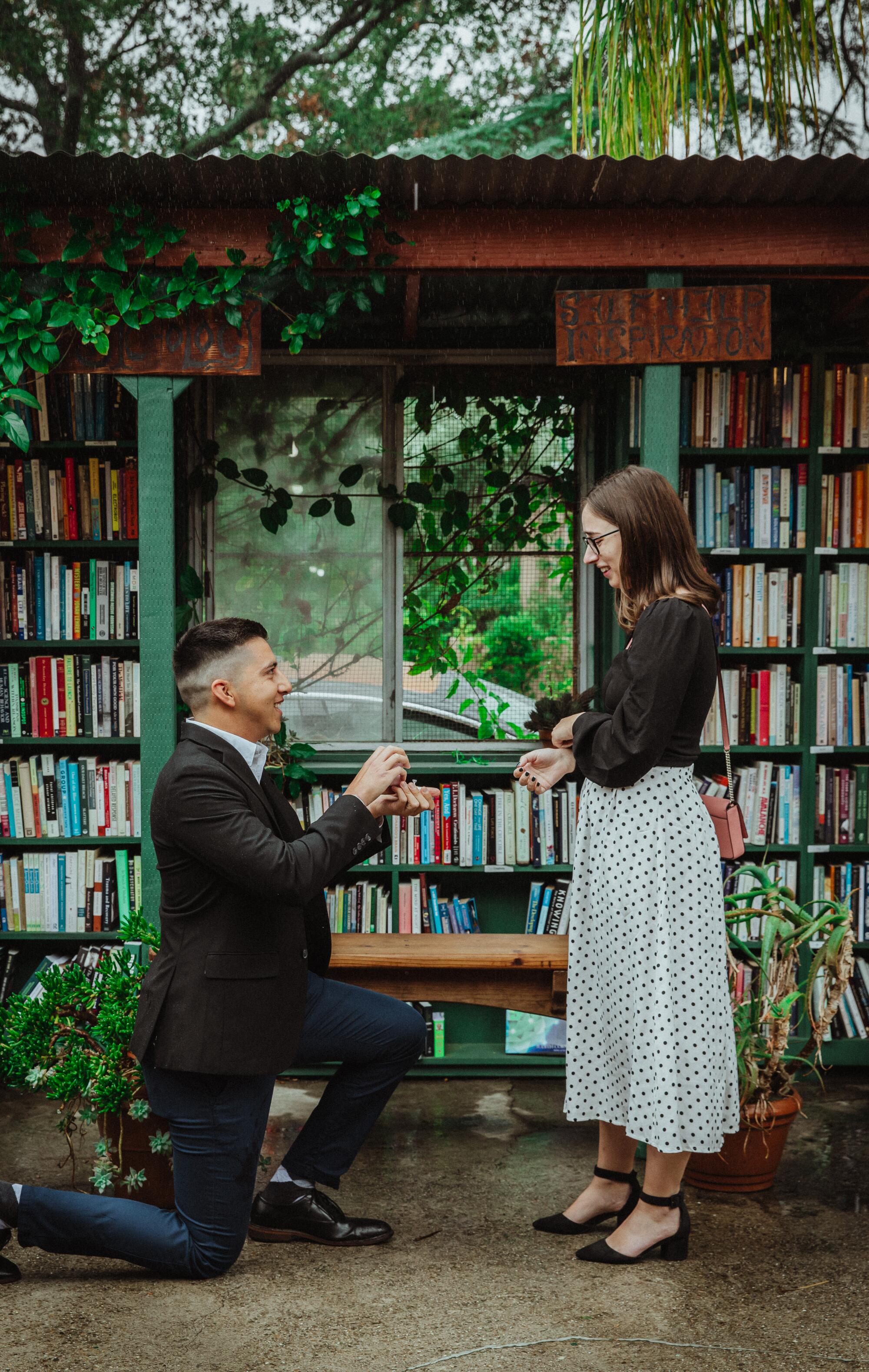 A man on one knee, holding a ring box, proposing to a woman in front of bookshelves at Bart's Books.