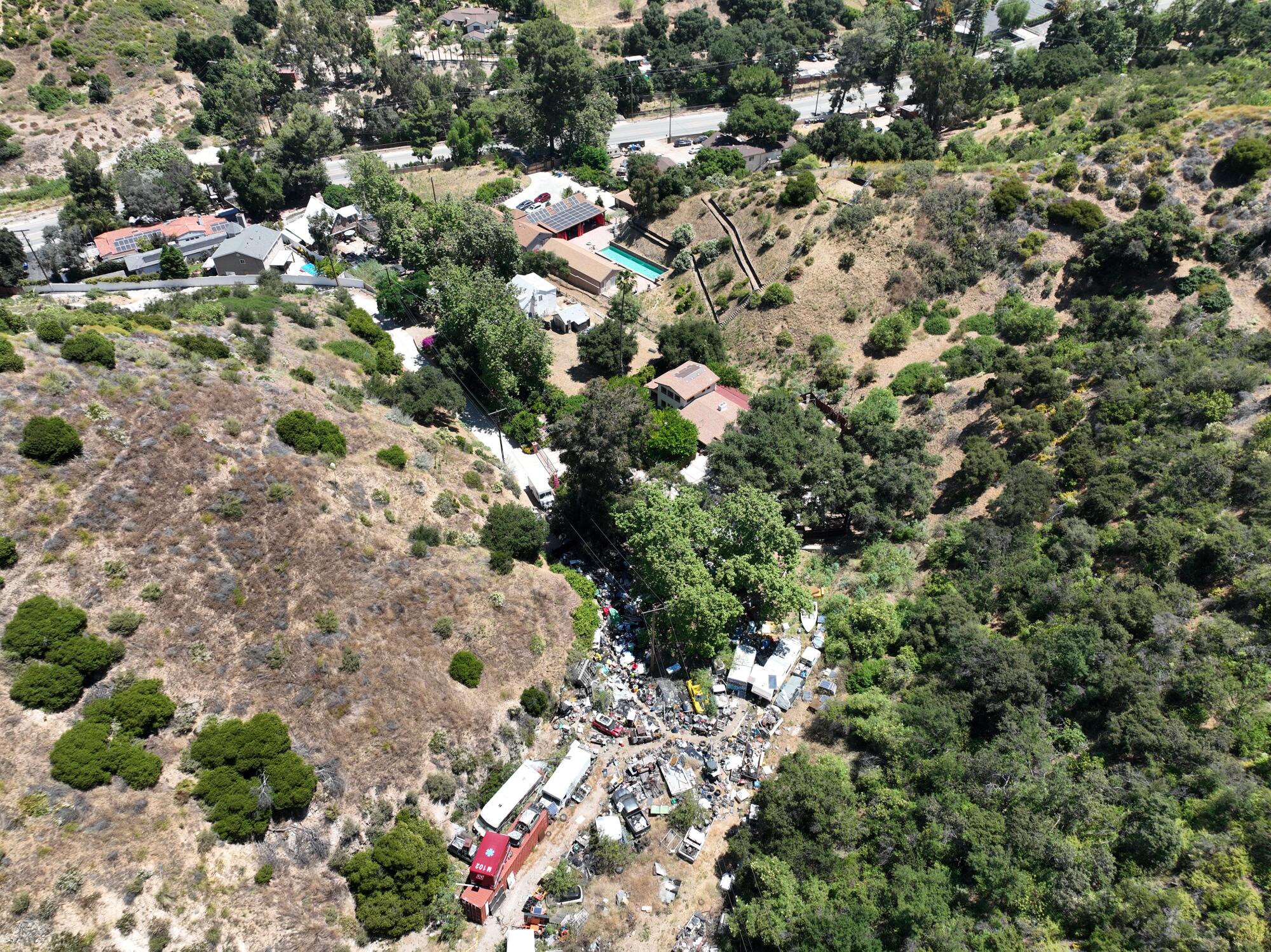 An aerial view of a rural property flanked by trees and grassy hills and crowded with vehicles.