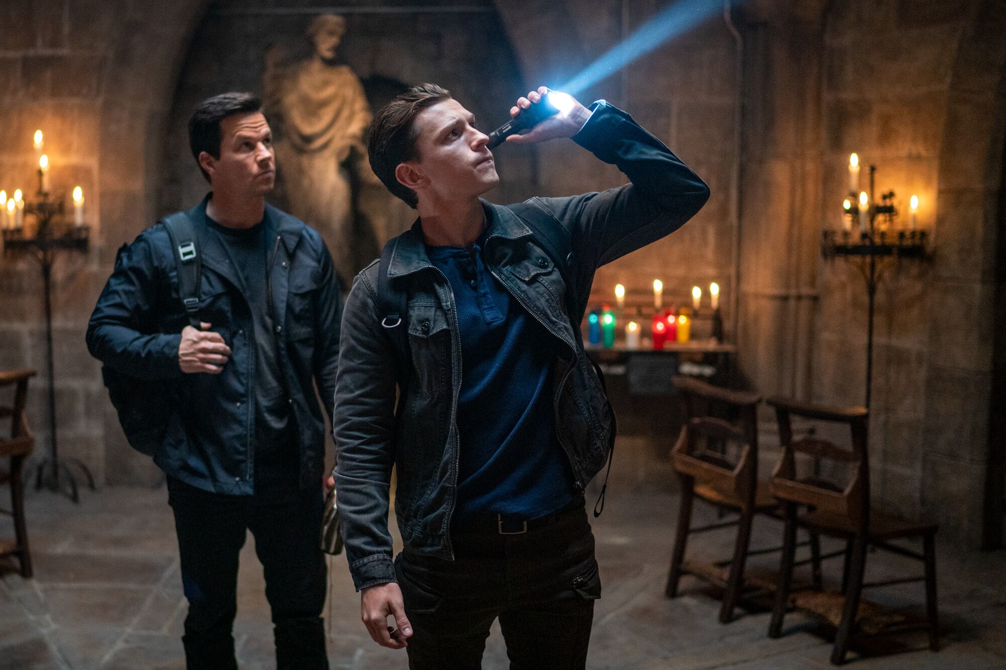 Two men in dark clothing stand amid lighted candles and votives in a religious space.