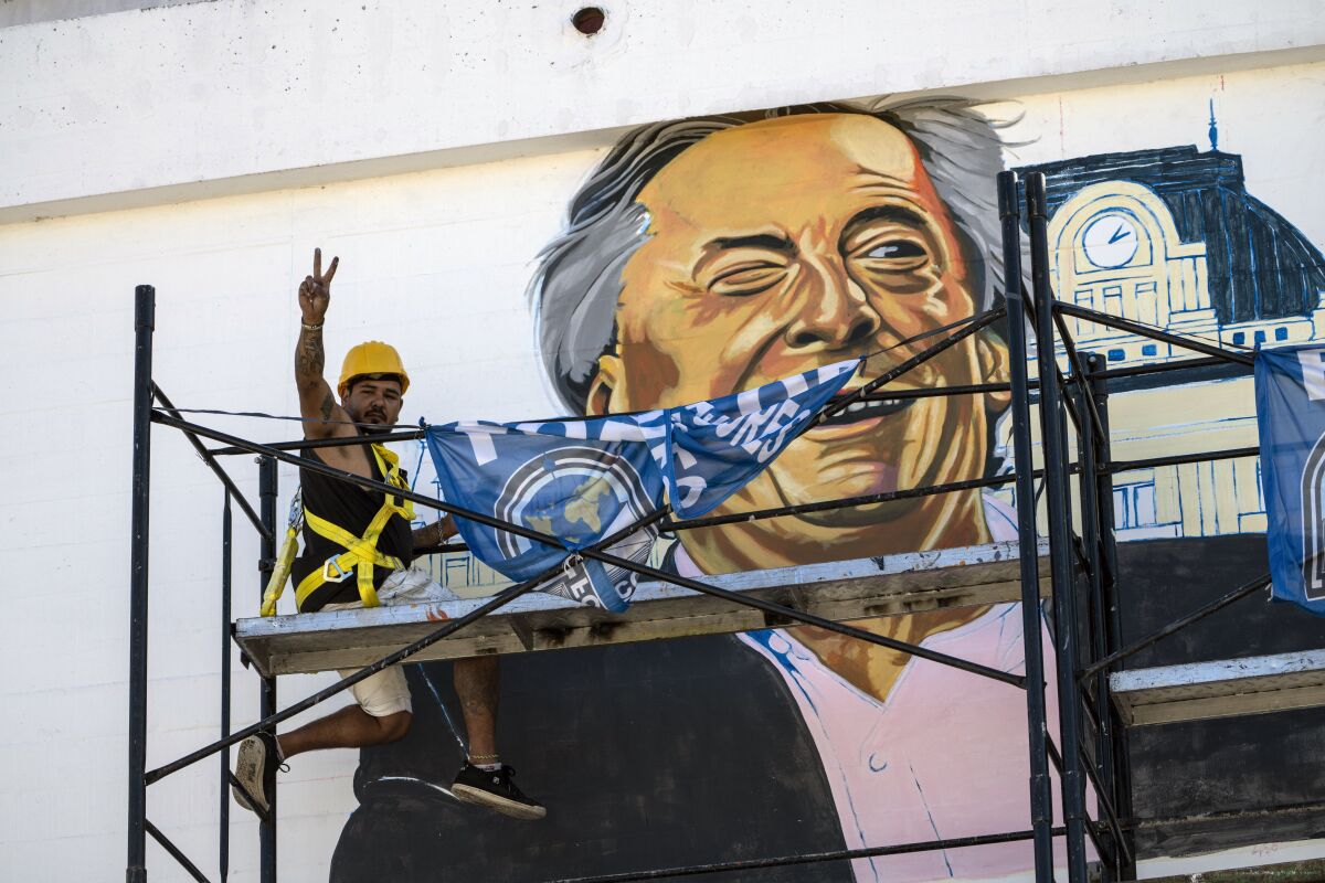 A worker flashes a victory sign, representing the Peronist Party, from a scaffolding as he paints a mural of former Argentine President Nestor Kirchner, the late husband of current Vice President Cristina Fernández de Kirchner, on a government building near the federal court in Buenos Aires, Argentina, Tuesday, Feb. 15, 2022. At the court, current Argentine President Alberto Fernandez made an unusual court appearance to defend his vice president — and former chief — calling allegations of corruption during her term as head of state “a sort of fantasy.” (AP Photo/Rodrigo Abd)