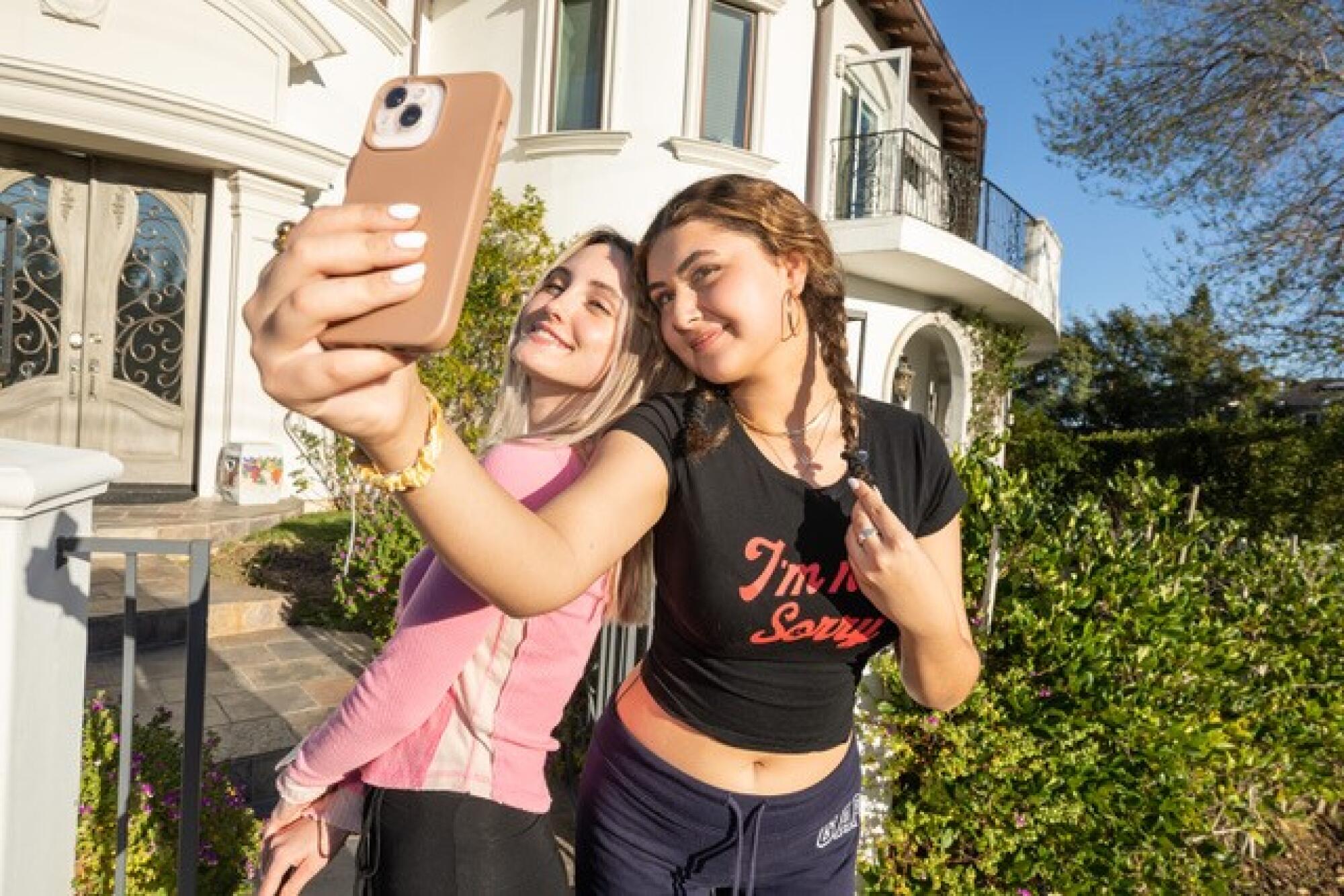 A blond girl in a pink top poses next to a girl in a black top who is holding a phone out in front of them.