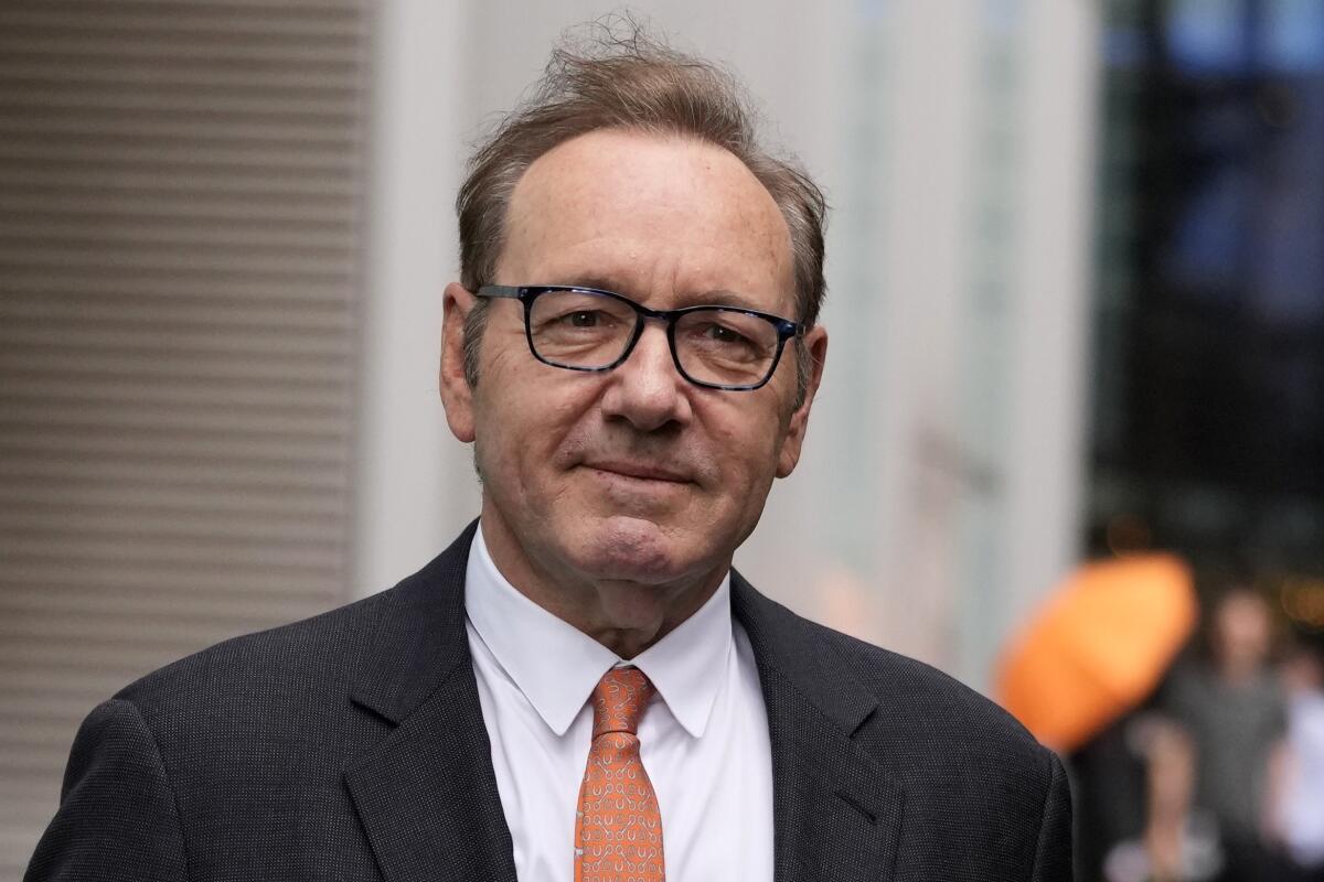 Kevin Spacey in a dark suit and orange tie outside a building