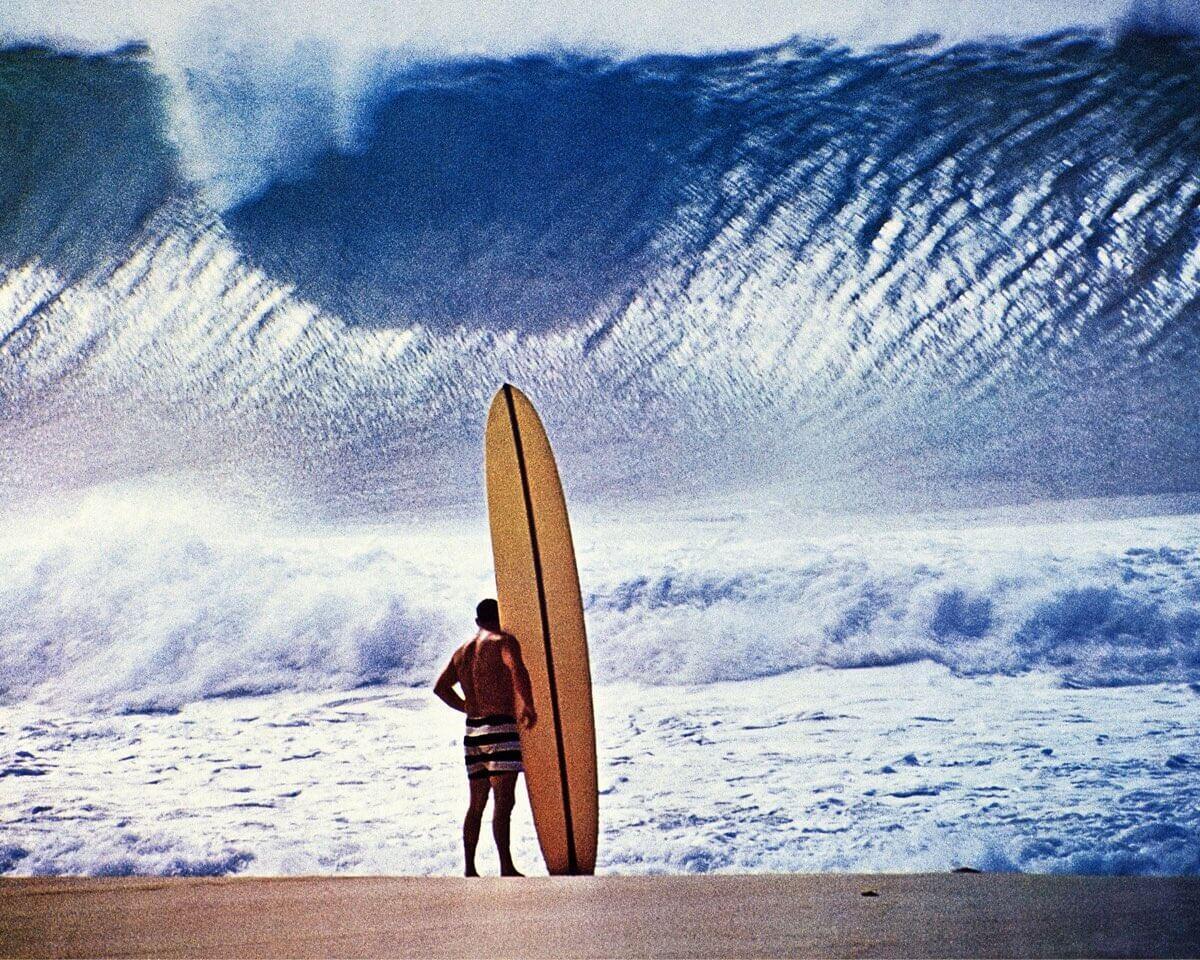 A man with a longboard faces a giant wave on a beach.