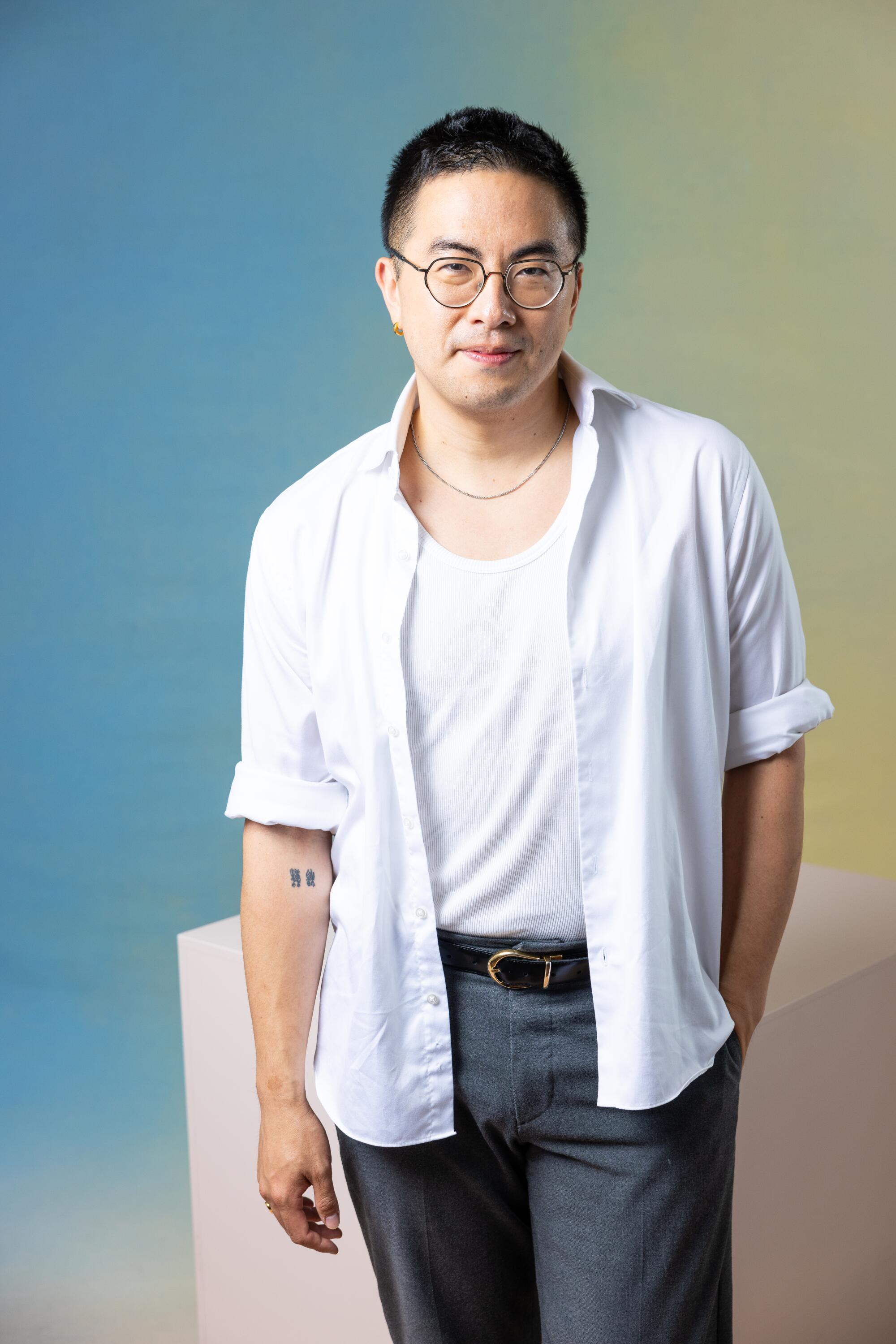 Bowen Yang stands in a white shirt and gray pants in front of a cube.