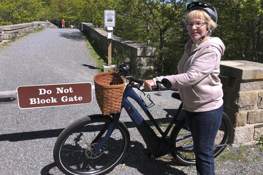 FILE-In this June 8, 2019 file photo, Janice Goodwin stands by her electric-assist bicycle at a gate near the start of the carriage path system where bikes such as her are banned inside Acadia National Park, in this photo June 8, 2018, in Bar Harbor, Maine. Interior Secretary David Bernhardt signed the order on Thursday allowing motorized electric bicycles into national parks and other public lands.(AP Photo/David Sharp, files)