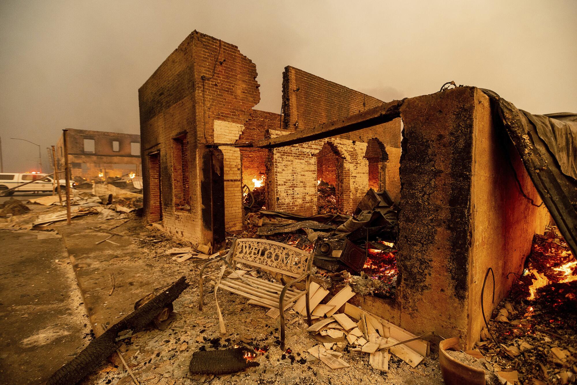 A sidewalk bench, its wrought iron "welcome" twisted by flames, stands outside a building gutted by fire