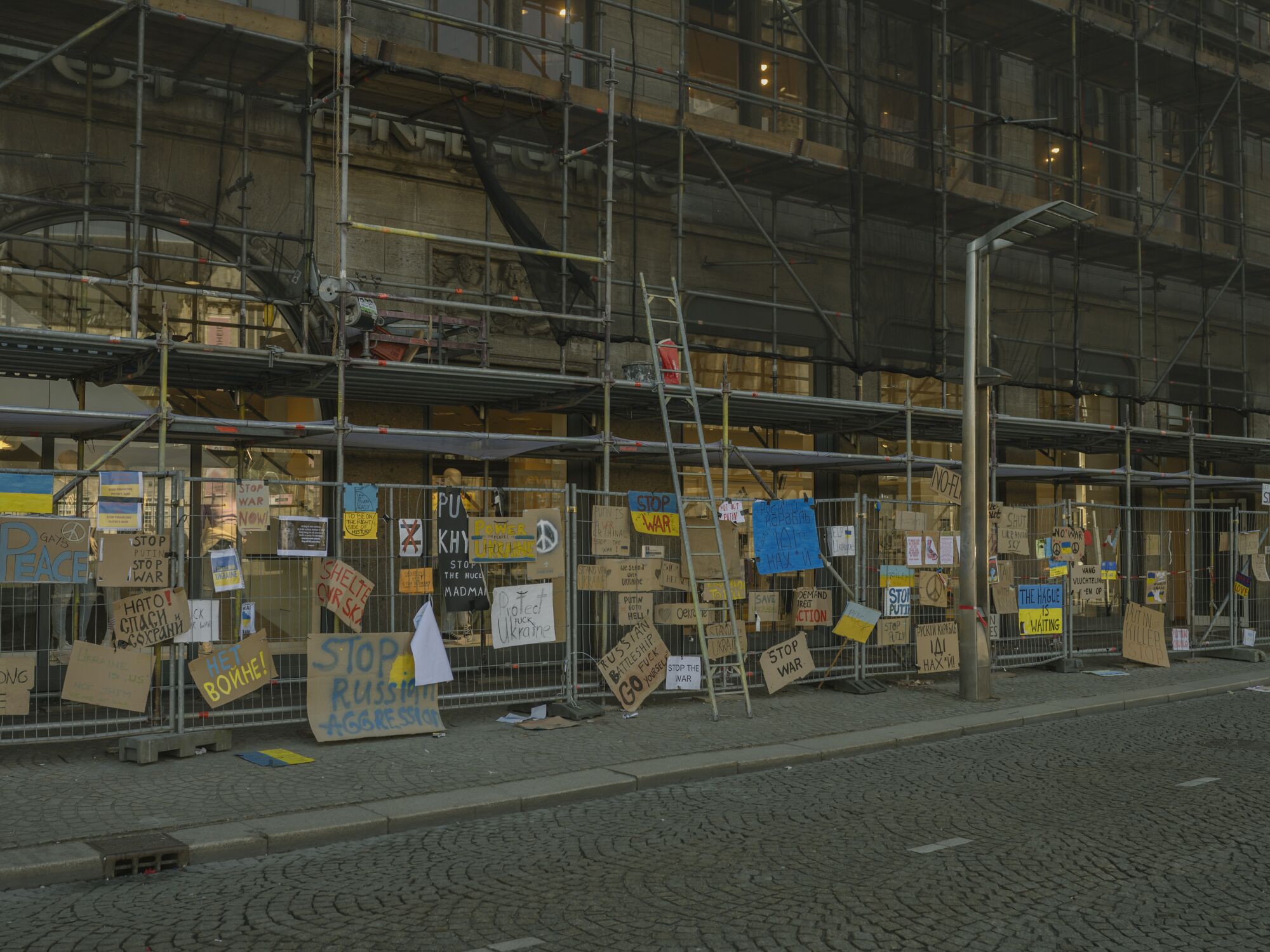 Many cardboard signs are hung on a fence below scaffolding 