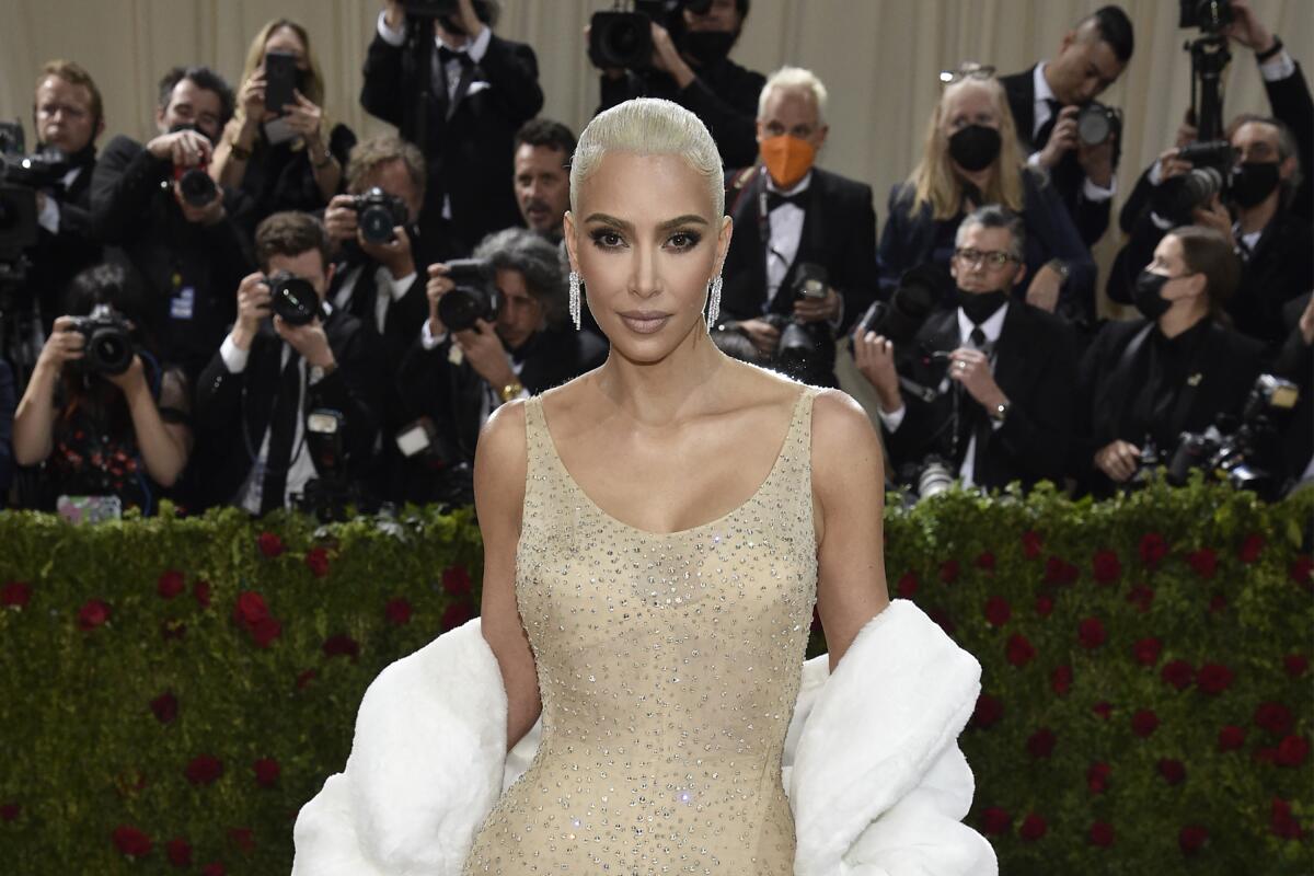 A blond woman with slicked-back hair wearing a jeweled gown and fur stole poses in front of photographers