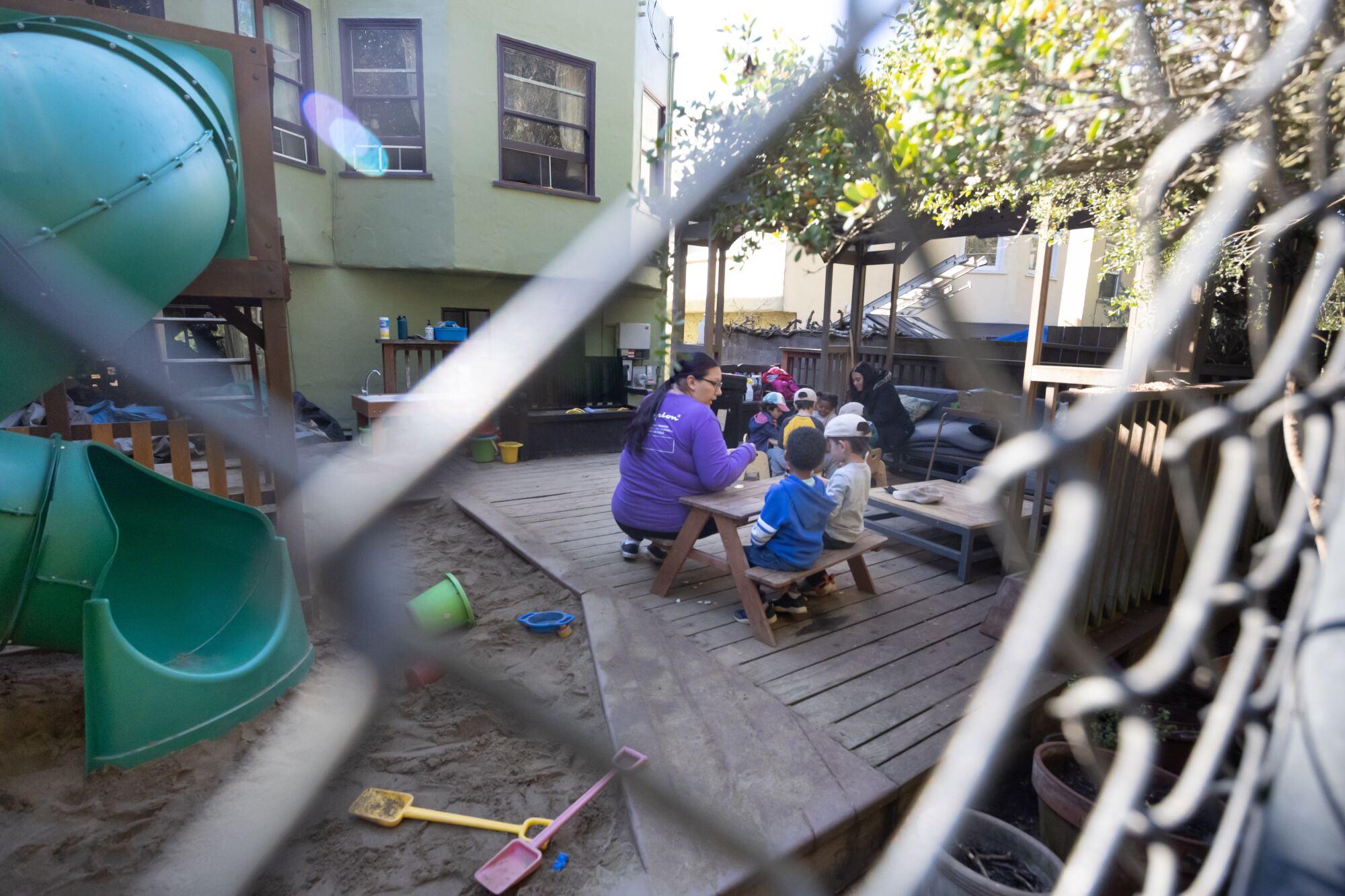 A view through a chain-link fence of two women seated at small picnic tables with several children in a leafy backyard.