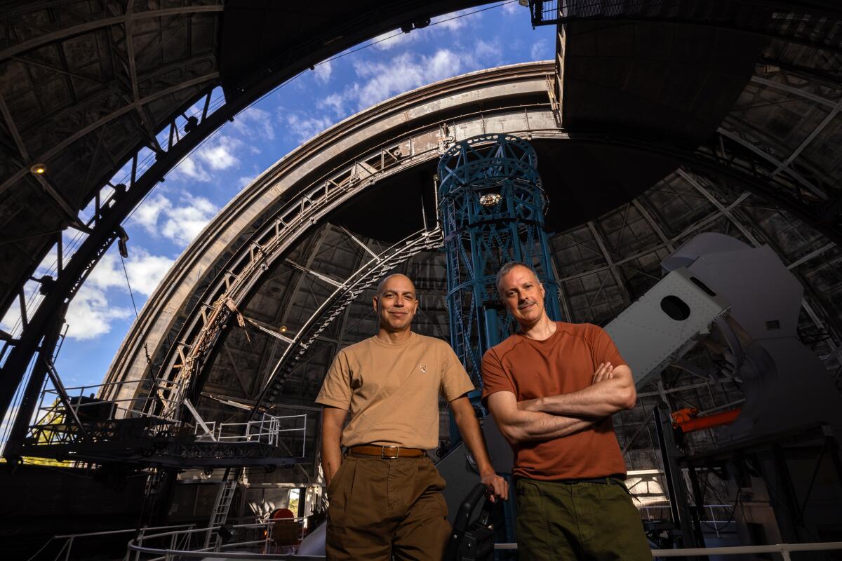 Two men stand on a walkway inside a domed observatory that has one of its ceiling panels open to the sky.