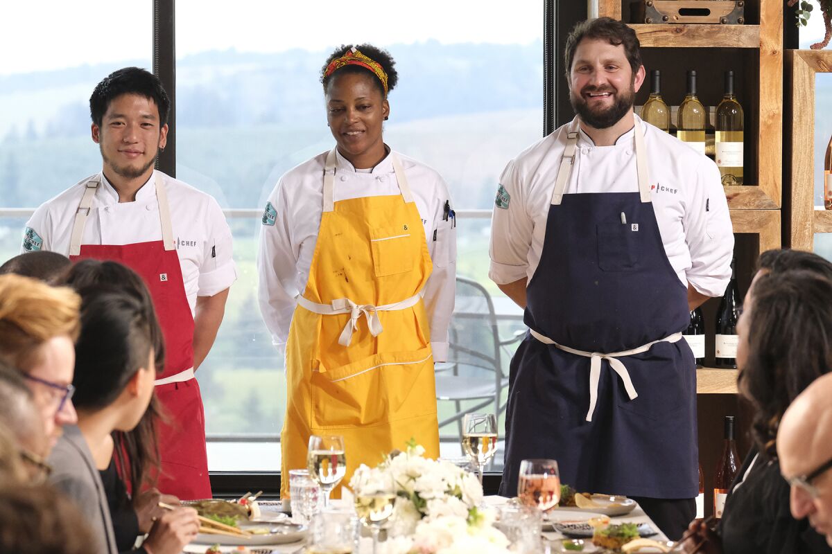 Three chefs stand in front of guests seated at a set table.