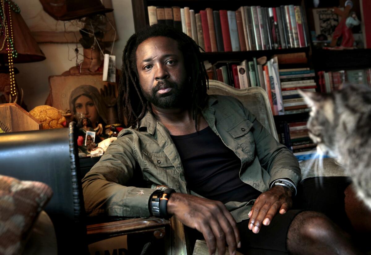 Marlon James poses for a photo in a room filled with knickknacks, books and a cat.
