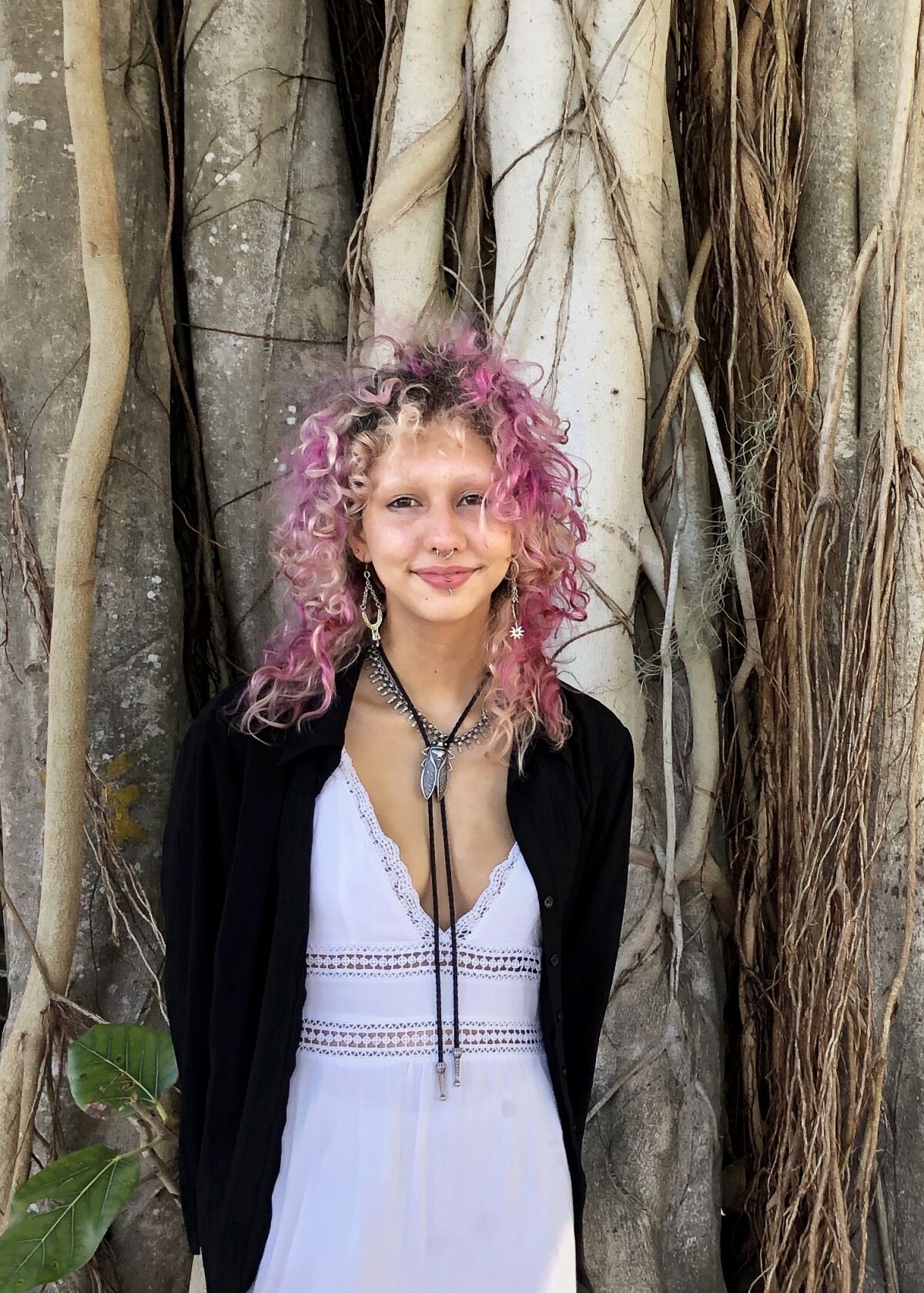 A young person with curly pink hair, wears a white dress and a black cardigan, smiles while posing in front of a large tree. 