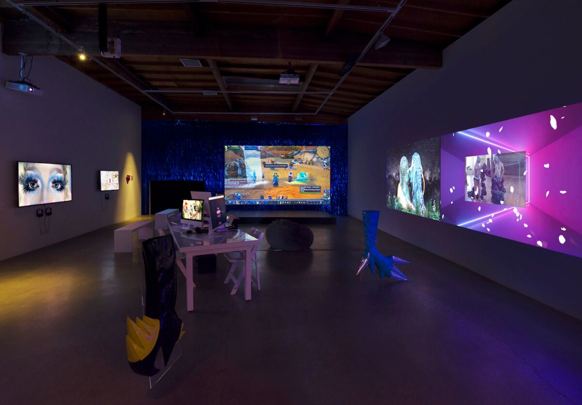 A darkened gallery features video on three walls — one bathed in purple light — with objects at center.