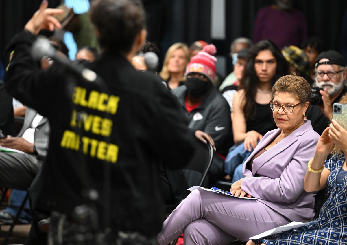 Mayor Karen Bass seated in crowd of people as an activist, standing, gestures while speaking