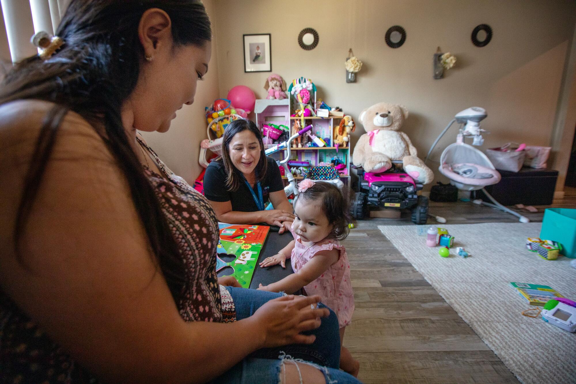 Two women play with a baby girl in a living room filled with toys. 