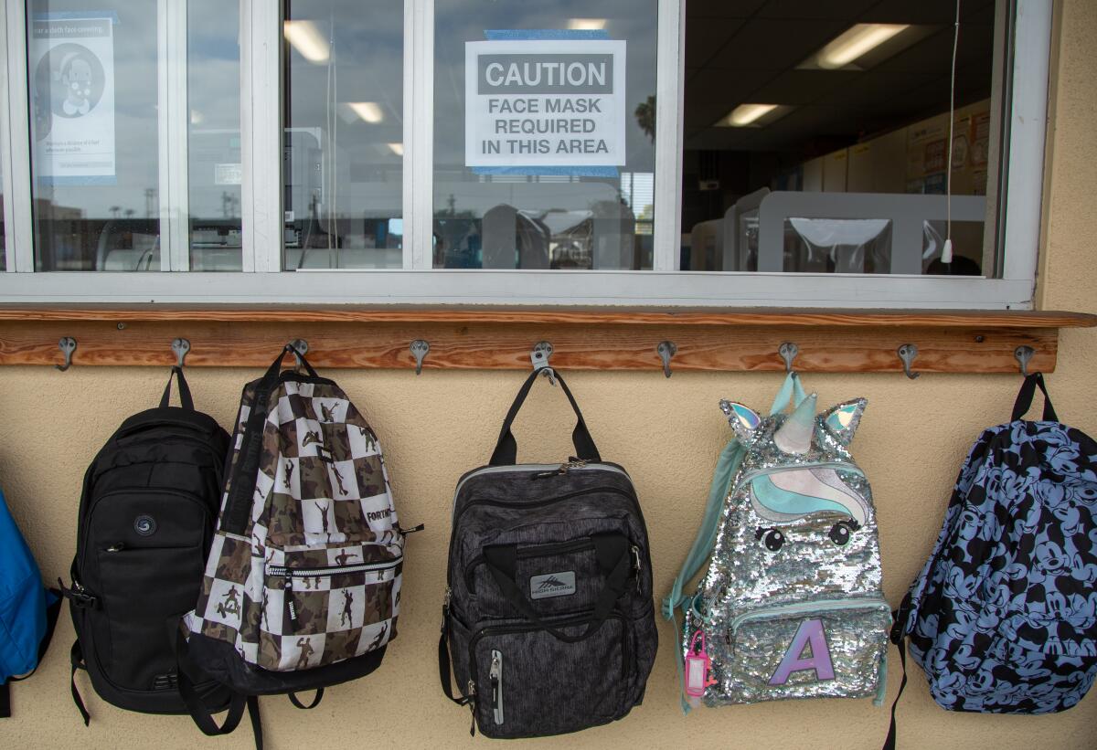 Backpacks hang on hooks, and a sign is taped to a classroom window: "Caution: Face mask required in this area"