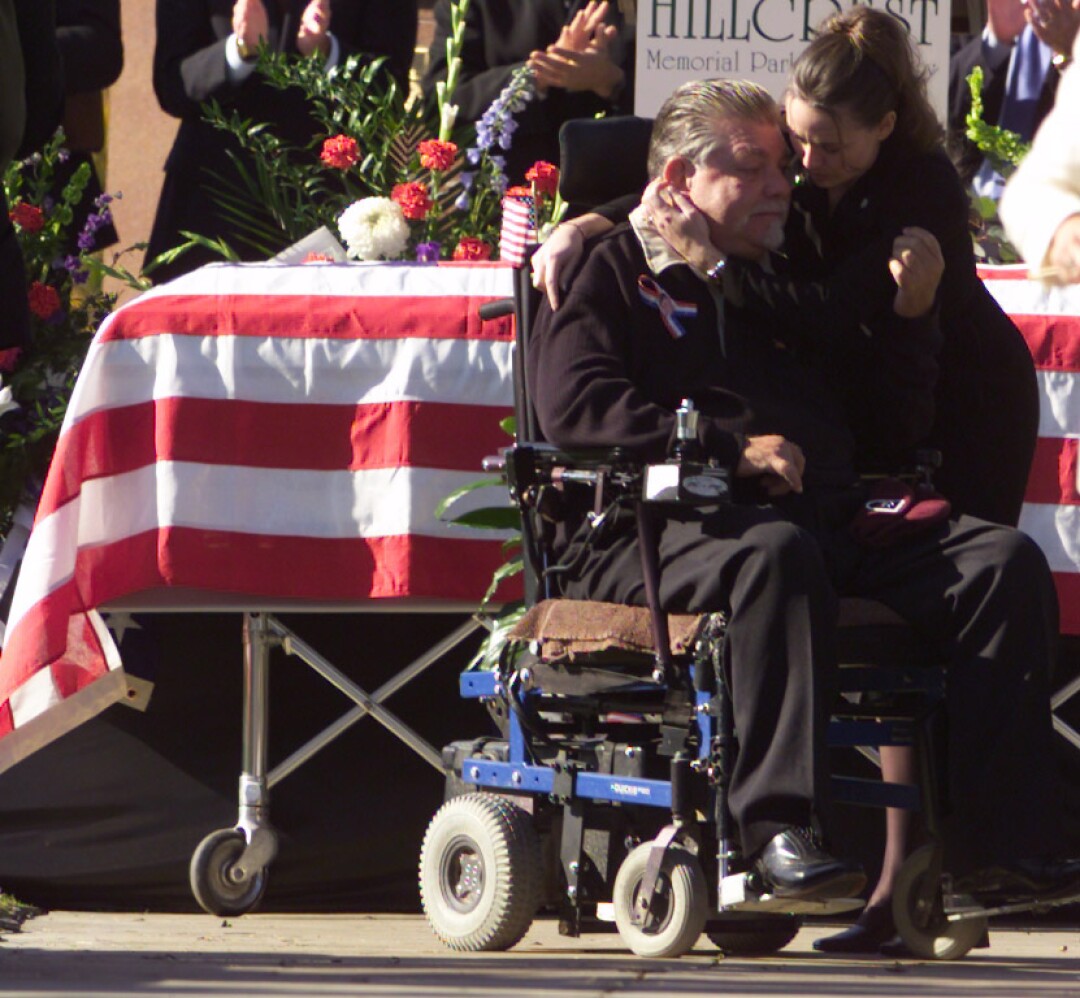 A man in a wheelchair and woman hug by a coffin.
