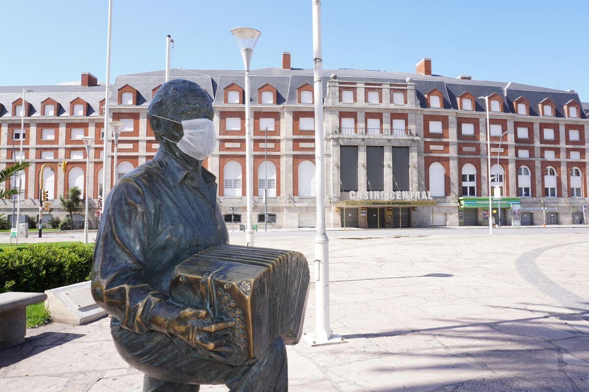 A bronze statue of a man playing a bandoneón, outside a stadium, is adorned with a face mask.