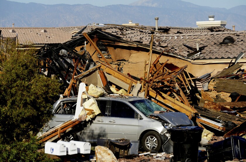 Charred, wrecked debris from a home explosion partially covers a minivan, with another damaged home in the background
