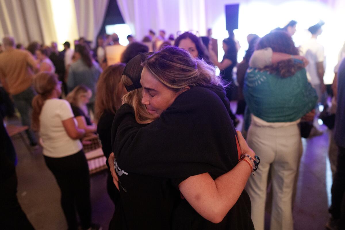 Two people dressed in dark clothes hug each other in a room full of people.