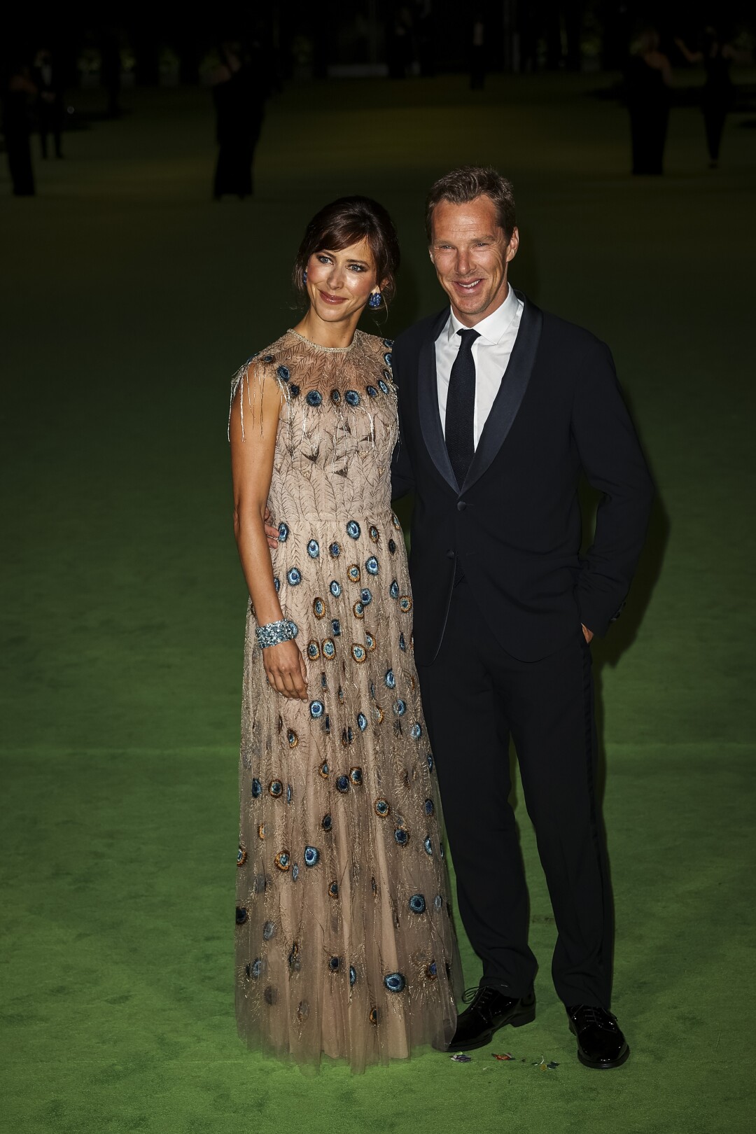 A woman in a patterned dress and a man in a black suit and tie posing on a green carpet