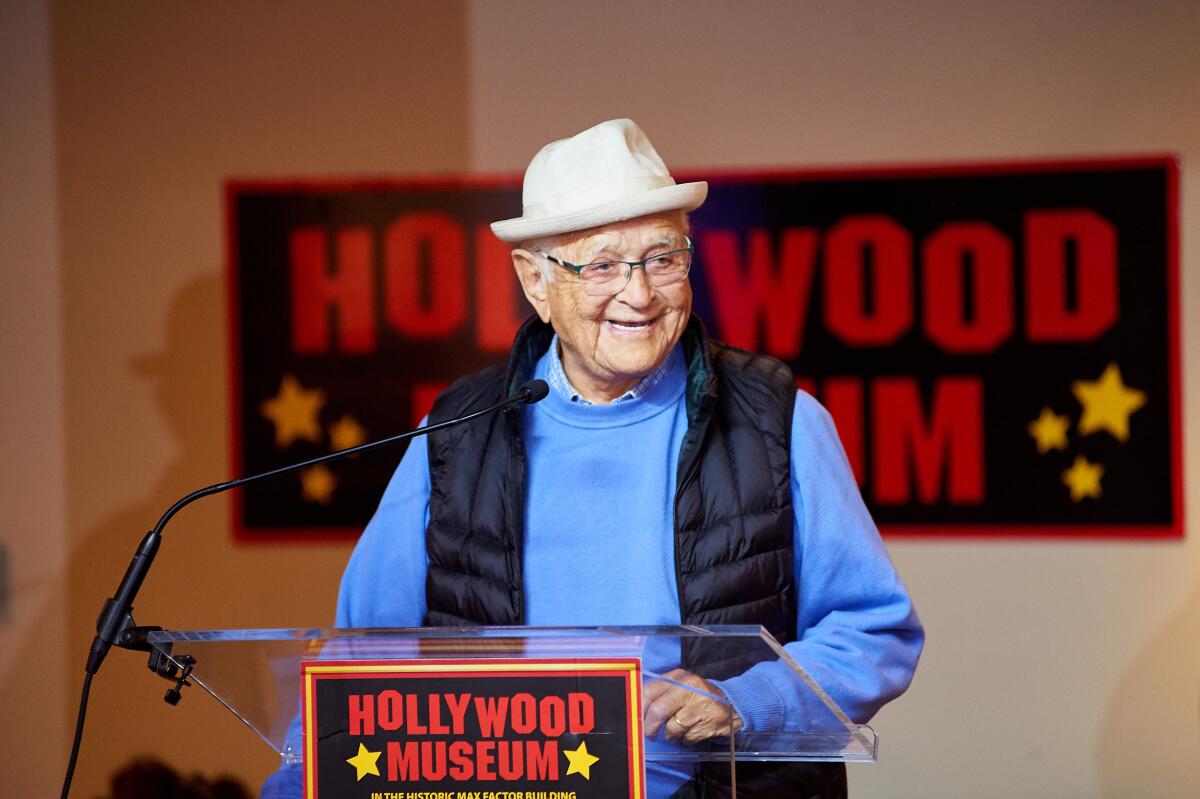 Norman Lear in a white hat, blue sweater and black puffer vest stands at lectern with a Hollywood Museum sign in front of it.