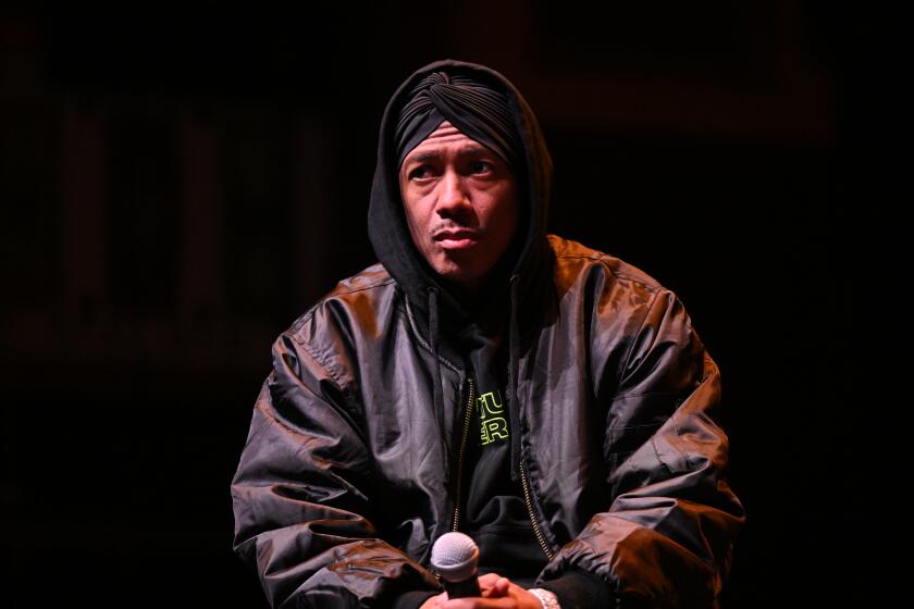 Nick Cannon sits onstage in a hoodie and leather jacket, holding a microphone on his lap with both hands.