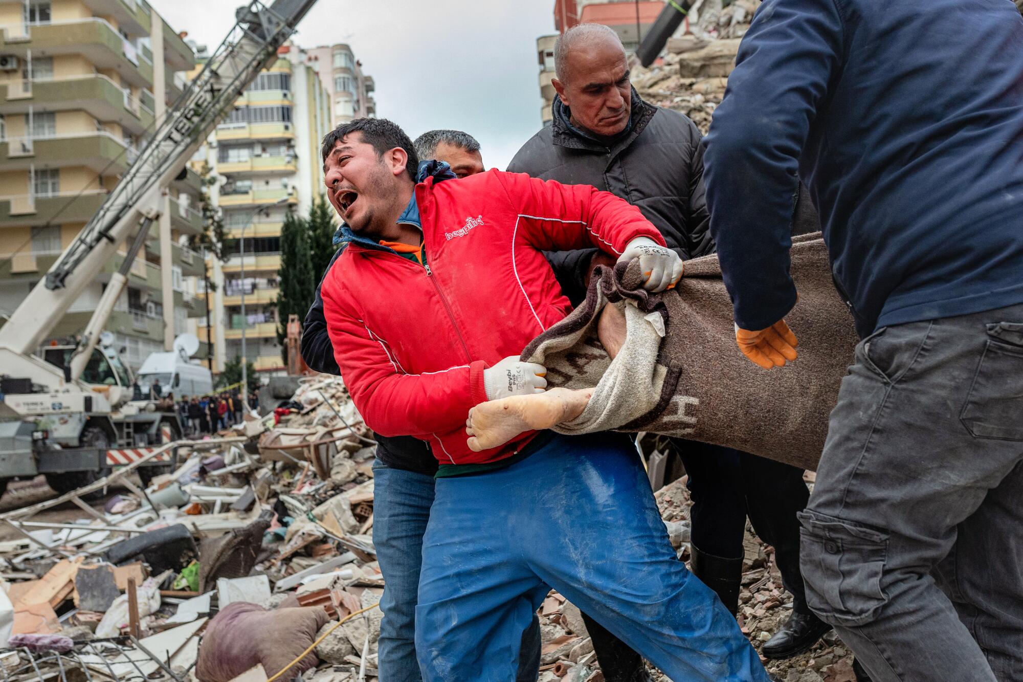 A rescuer cries out as he helps carry a bod wrapped in blanket, with a human foot sticking out.
