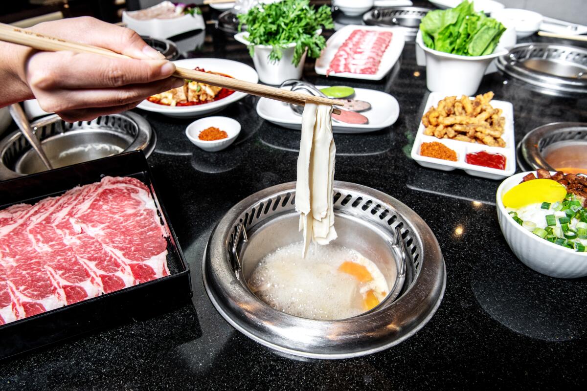 A diner cooks noodles in hot broth in a metal pot on a table full of sliced raw meat, vegetables and other dishes