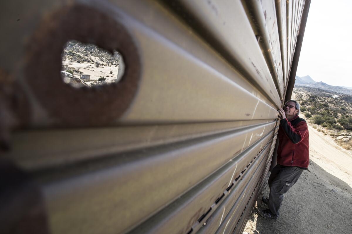 Bill Pape peers through a border fence made of Vietnam-era steel airstrip landing mat near his home in Jacumba Hot Springs, Calif.