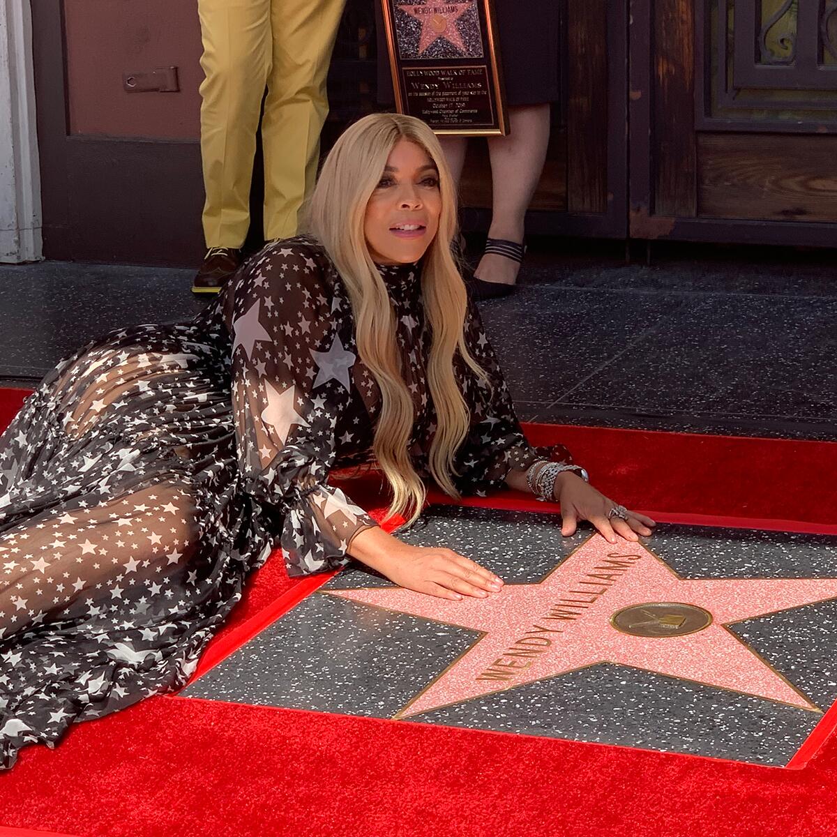 Wendy Williams in a black dress with white stars, reclines next to her star on the Hollywood Walk of Fame.