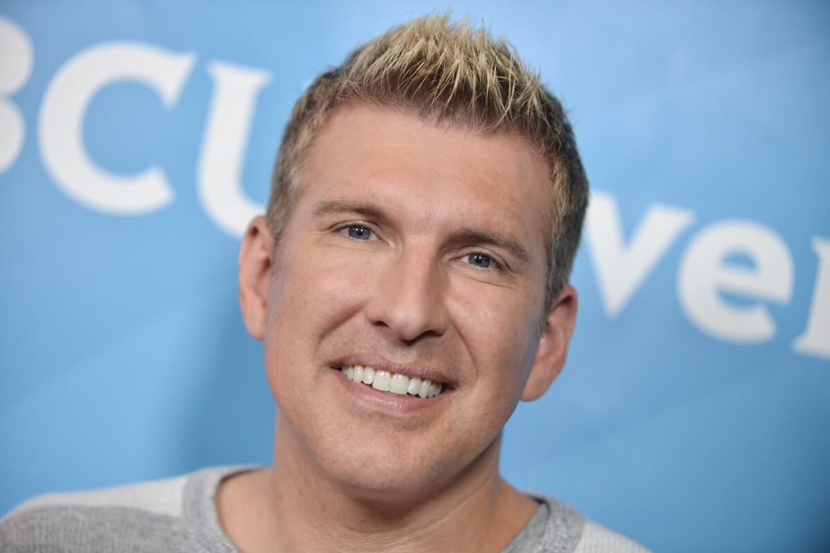 A smiling Todd Chrisley wears a striped shirt and stands against a light blue backdrop.