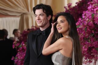 John Mulaney in a black suit posing next to Olivia Munn in a silver gown at the Oscars red carpet