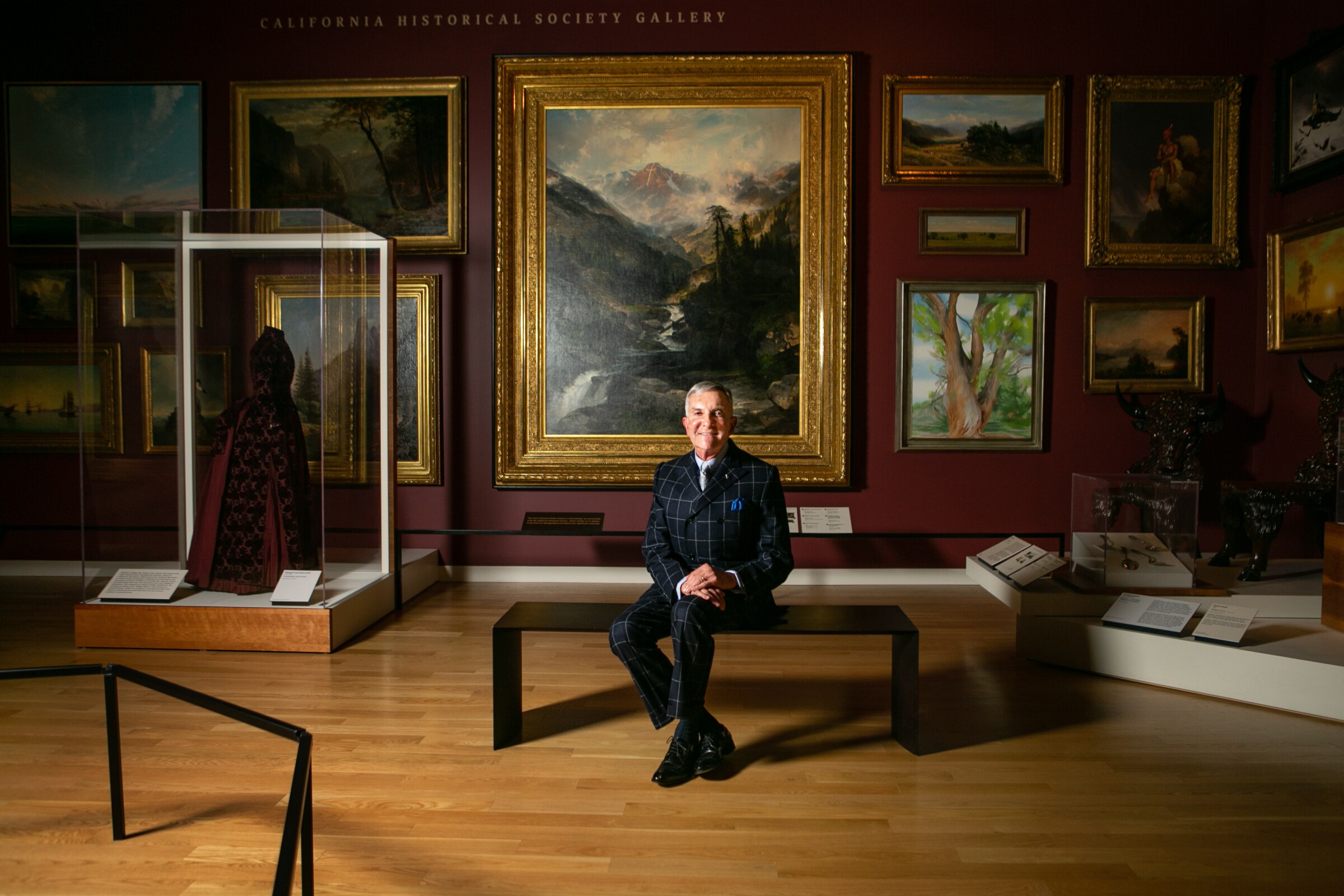 Rick West, in a black and blue checked suit, sits before landscapes in a dramatically illuminated gallery