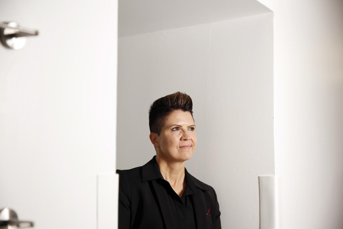 A woman wearing a dark top and jacket stands in an alcove between two white walls.