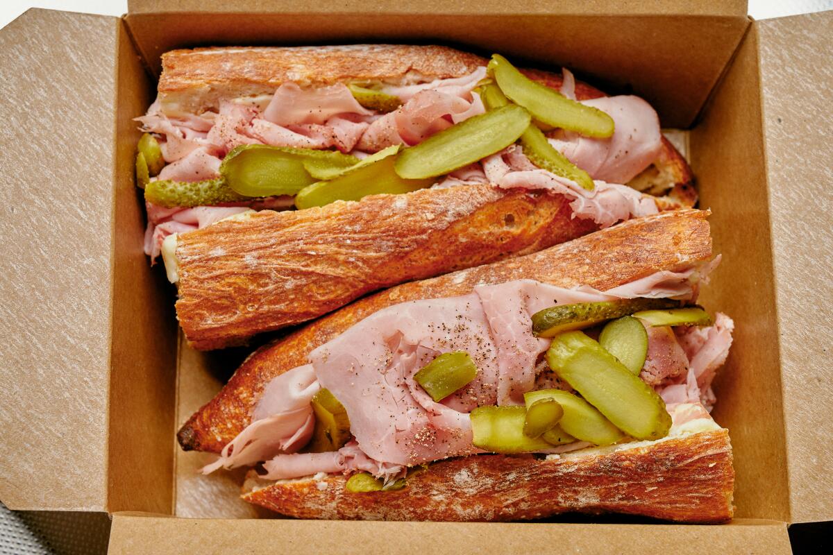 Two halves of a jambon beurre sandwich with cornichons in a cardboard takeout container