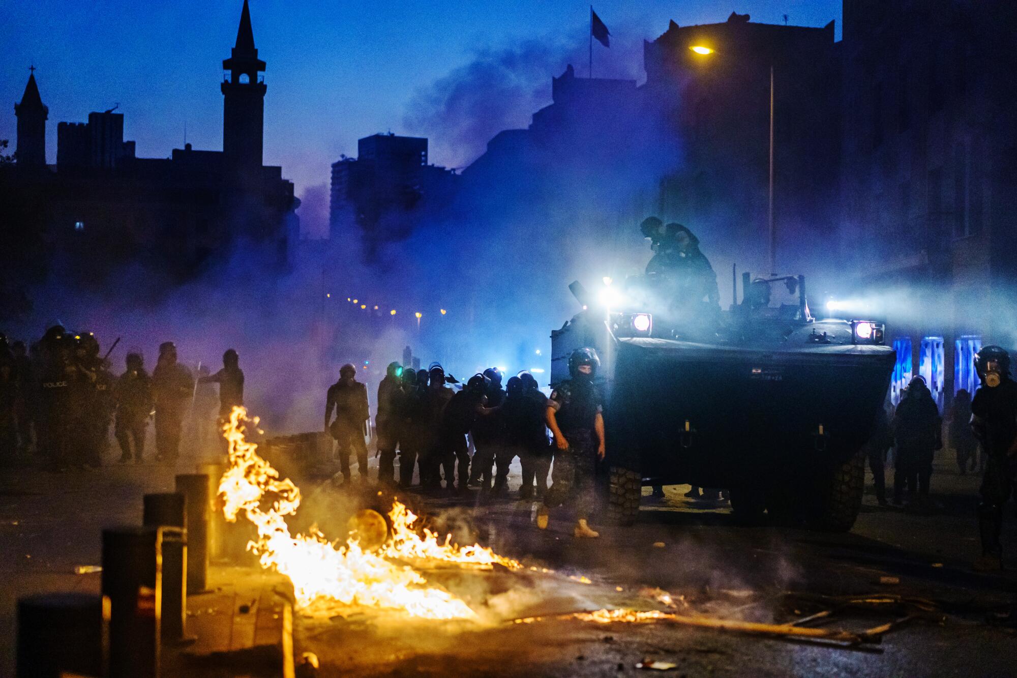 A police vehicle and officers in riot gear move through purplish haze and past flames leaping on the street.